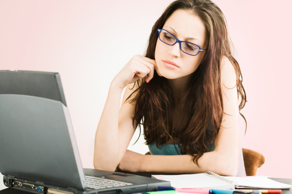 A woman sits at a computer, and looks at the screen, lost in thoughts. We can see exercise book on her table, and she wears glasses