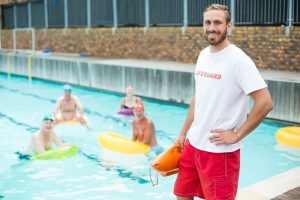 Man is doing his job of a lifeguard. We can see him standing at a side of a swimming pool, in a lifeguard t-shirt