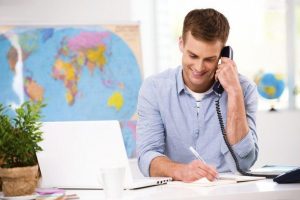 Travel agent smiles when talking to the client on the phone. We can see a latop, map of the world, and other things in their office.